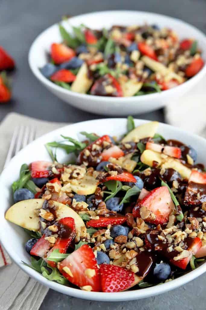 This is a picture of a mixed green salad topped with fresh sliced apples, strawberries, blueberries and nuts. Along with a chocolate vinaigrette.