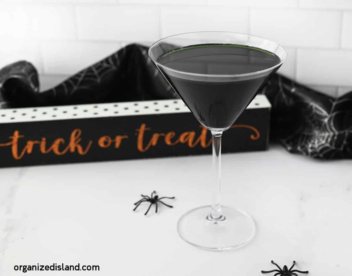 A martini glass filled with a black beverage sits on a white table. Nearby are small, plastic spiders and a black cloth with spiderweb patterns. In the background, a sign reads "trick or treat" in orange letters. The URL "organizedisland.com" is visible in the bottom left corner.