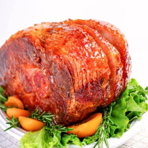 Juicy glazed ham on a white plate with fresh greens and orange slices.