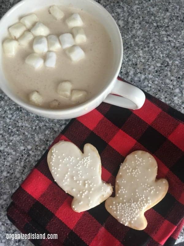 Top down view of sugar cookies shaped like mittens on a red and black buffalo print napkin, next to a mug of hot chocolate.